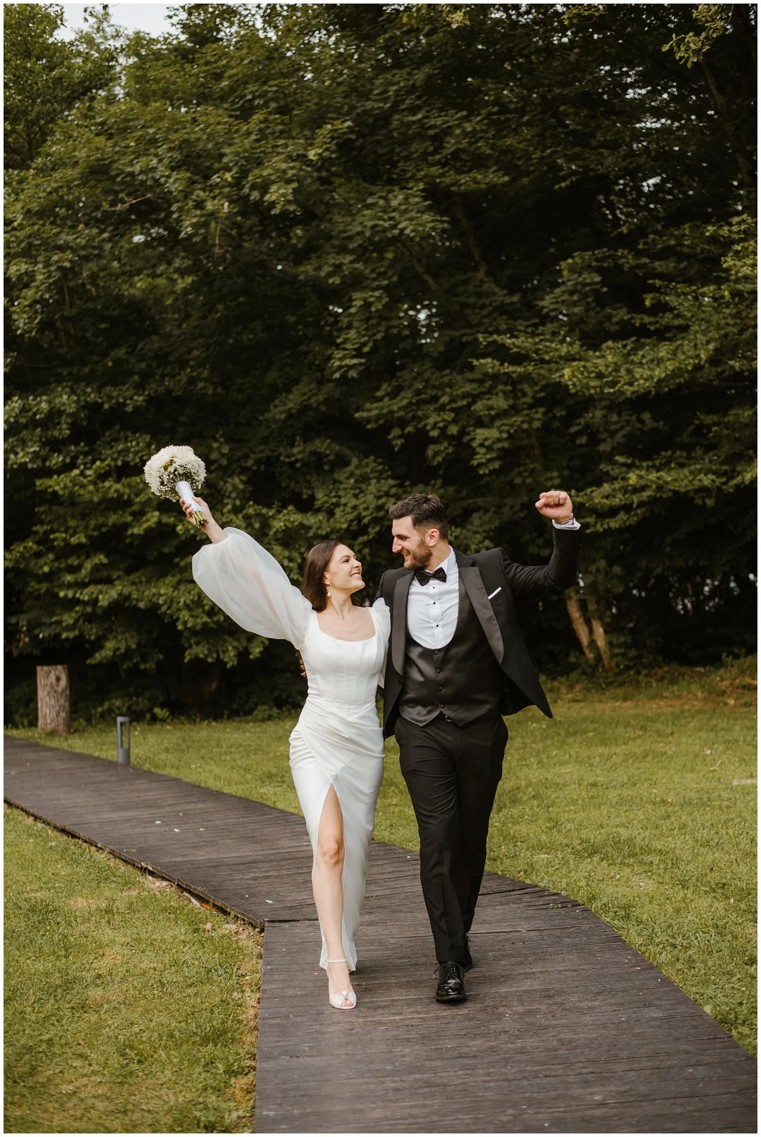 Mladenci hodaju drvenom stazom, ona podiže buket, oboje uzdignutih ruku, okruženi zelenilom u Corberon Wedding Resortu